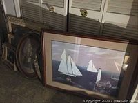 Nine pieces of wall art leaning against a wall including a large nautical sailboat print with a lighthouse visible.