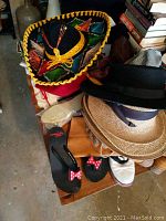 Photo showing assorted hats including a colorful sombrero, woven straw hats, black felt hat, black slippers with red polka dot bows, flip flops, brown boots and white slip-ons on a wooden surface with books in background.