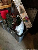 Photo of an open gray plastic trunk filled with holiday decorations including a boxed fiber optic Christmas tree, several small artificial wreaths with pine cones, and green placemats.