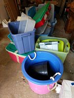 Photo showing various plastic totes including large round bucket style with rope handles in pink and blue, plastic crates, and rectangular totes in multiple colors.