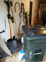 Large green outdoor trash bin showing scuff marks and lid, with containers of garden chemicals next to it.