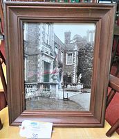 Framed black and white photograph of an old brick building with bird sculpture on pedestal visible outside. Wood frame has stepped design and medium brown finish.