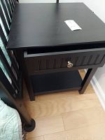Black wooden side table viewed from a corner angle showing rectangular top, drawer with vertical grooved design, and lower shelf.
