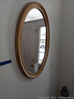 Oval mirror mounted on a white wall reflecting a door and window, showing gold frame detail and condition.