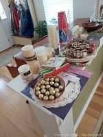 Photo showing various candles, decorative doilies, paper napkins, plates, hanging decorations, and gold decorative balls arranged on a table in a well-lit room.