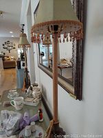 Photo showing the full length of one table lamp with metal base and tasseled beaded jewel shade, on a table near a mirror and household items.