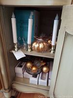 Shelf with two electric candles, blue boxes of crystal champagne flutes, gold pumpkin decoration, crystal bowl with gold ornaments and packaging boxes.