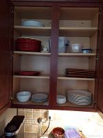 Inside of wooden kitchen cabinet showing stacks of red, white, and striped plastic plates with some plastic storage containers and a butter dish.