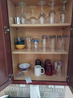 Three shelves inside a cabinet with assorted glassware including stemmed wine glasses on top shelf, various clear glasses on middle shelf, and mugs and bowls on bottom shelf.
