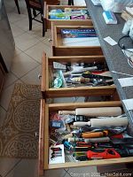 Four open wooden kitchen drawers filled with assorted kitchen utensils and supplies including rolling pins, knives, and miscellaneous tools.