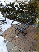 Two metal swivel chairs on a stone patio covered partly with snow, showing the full chair with round swivel base and curved backrest with circular decoration.