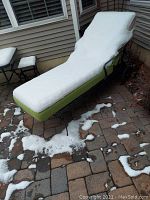 Side angle view of metal chaise lounge with green cushions covered partially by snow, showing the wheels and frame details.