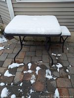 Square metal framed patio table with glass top covered in snow showing table surface and legs on brick patio.