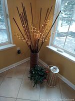 Tall lighted artificial plant in woven basket next to small artificial flowering plant and white lighthouse candle holder on floor by windows.