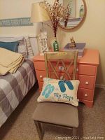 Room corner with wooden chair holding a pillow with flip flop design, bedside dresser with orange drawers, and a round rope framed mirror on the wall.