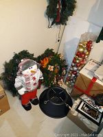 Photo showing two Christmas wreaths, snowman figurine, black metal tree stand, and plastic container with Christmas ornaments.
