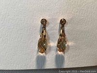 Pair of Swarovski earrings shown from the front hanging vertically. Each has a small round crystal stud on top with a faceted teardrop crystal pendant in golden shadow color.