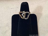 Front view of Swarovski ring showing double interlocked hearts, one gold and one crystal-encrusted, positioned on a black display stand.