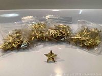 Five sealed plastic bags each containing gold star pin badges arranged in a row with one gold star pin badge placed in front of them on a white surface.