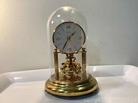 Clock under glass dome on brass base with white clock face and rotary pendulum.