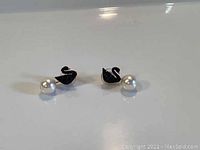 Pair of Swarovski earring jackets featuring black swan motifs and white pearl pendants on a white reflective surface.