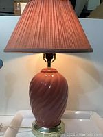 Front view of the lamp illuminated, showing the peach pink ceramic base with spiral swirl pattern and plaited lampshade.