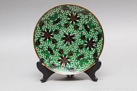 Front view of cloisonné enamel footed plate on display stand, showing green background with black and white floral and scrollwork pattern with gold outlines.