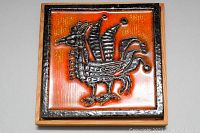 Front view of the ceramic tile showing the raised bird design on an orange background inside a detached wooden frame.