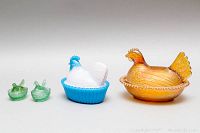 Front view of all five glass chicken-shaped items: two small green salt cellars, one medium blue and white milk glass bowl, one large amber Depression glass bowl.