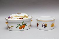 Photo of two casserole dishes side by side, showing fruit and wheat designs on oval covered dish and fruit on round dish