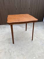 Front view of square folding card table with scalloped edges and cherry wood finish, standing on concrete floor.