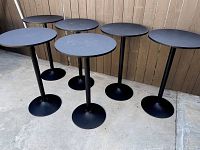 Six black skinny round cocktail tables positioned on concrete floor next to wooden fence.