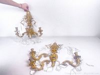 Two ornate gold-tone wall sconces with scrollwork detail and clear crystal prisms shown resting on a surface, one is broken into multiple pieces