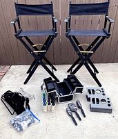 Two black director's chairs with fabric seats and backs, brass brackets, makeup box, hangers, caster wheels, lint rollers, organizer holders and hairbrushes arranged on concrete