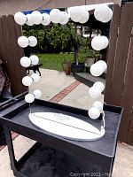 Square lighted make-up mirror with white bulbs around the edges, placed outdoors on a black cart.