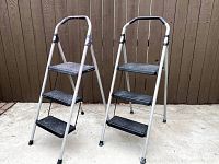 Two white metal step stools each with three black plastic steps, standing on concrete with a wood fence background