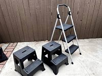 Three black plastic step stools and white metal folding ladder grouped together on concrete floor with wooden fence background