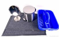 Front view of lot showing two black floor mats underneath small stainless steel trash can, blue plastic cleaning caddy, and round tabletop mirror.