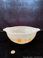 Side view of Pyrex Cinderella Bowl showing white milk glass with gold and orange floral-geometric pattern and dual handles, size compared to quarter coin.