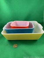 Three nested Pyrex refrigerator dishes in yellow, blue, and red with a coin for size comparison against green background.