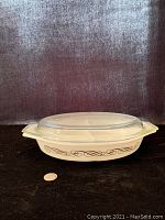 Side view of the Pyrex divided casserole with clear lid showing the black scroll pattern on the white glass body, placed beside a coin for size reference.