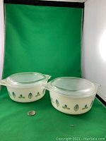 Two Pyrex milk glass servers with leaf pattern and lids on green backdrop with quarter for scale