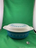 Side view of the assembled Pyrex casserole dish with lid, showing the blue snowflake pattern and lid design, placed against green backdrop with quarter for scale.