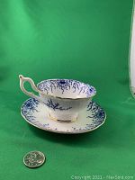 Side view of the blue wide mouth cup and saucer showing cobalt blue pattern and gold trim with coin for scale.