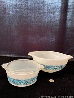 Pyrex Blue Horizon Cinderella mixing bowl and a round casserole with lid side by side on black background with coin for size reference.