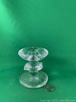 Clear textured glass Iittala Festivo 1 ring candlestick with a red label, next to a US quarter for scale, photographed against a green background from the side.