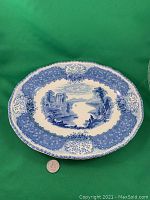 Top-down photo of blue and white Royal Doulton ceramic platter on green backdrop with coin for scale, showing scenic transfer ware design of ruins, water and nature.