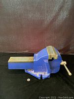 Side angle photo of blue mechanics vise showing the entire tool on a plain black surface with quarter coin for scale.