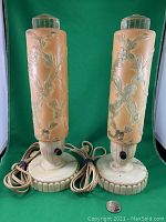 Pair of beige Mid-Century Modern textured glass bedside lamps with raised floral design, plastic ribbed bases, black rotary switches, and clear ribbed glass caps, shown against green background.