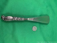 Full view of the vintage shoe horn, showing the entire length including the ornate silver handle and metal blade alongside a quarter for scale.